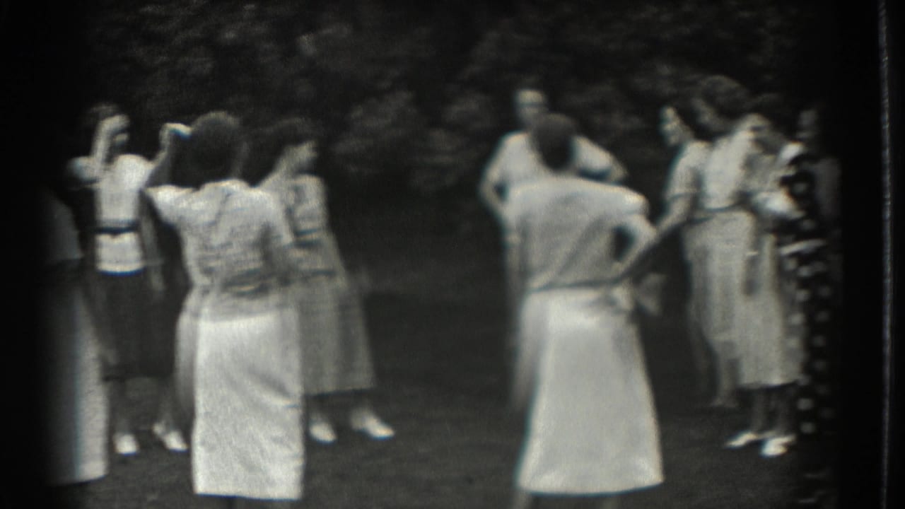 Women in 1930s summer clothing stand and gesture in a loose circle during a lawn game in Southampton.