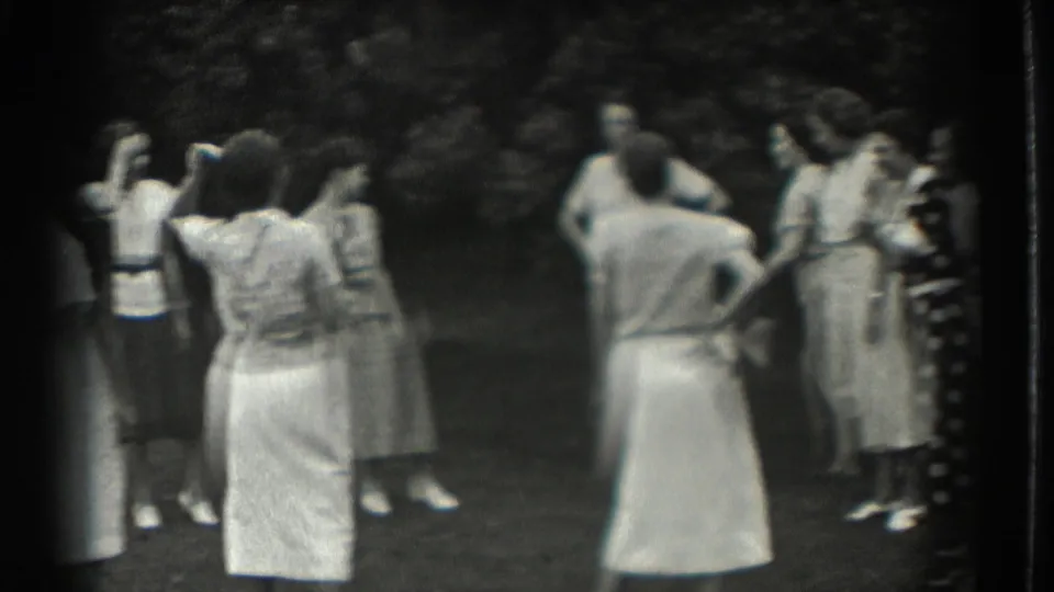 Women in 1930s summer clothing stand and gesture in a loose circle during a lawn game in Southampton.