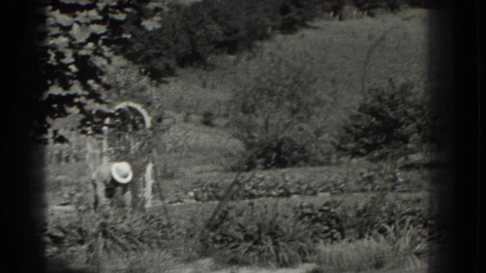 1937 Southampton home-movie frame showing a distant person near a white garden arch with rows of trees and shrubs behind.