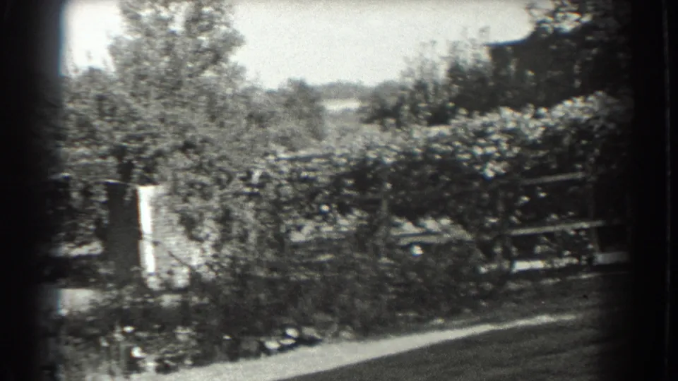 Black-and-white 8mm frame from Southampton showing trees, shrubs, fence sections, lawn, and pale hanging fabric near the garden edge.