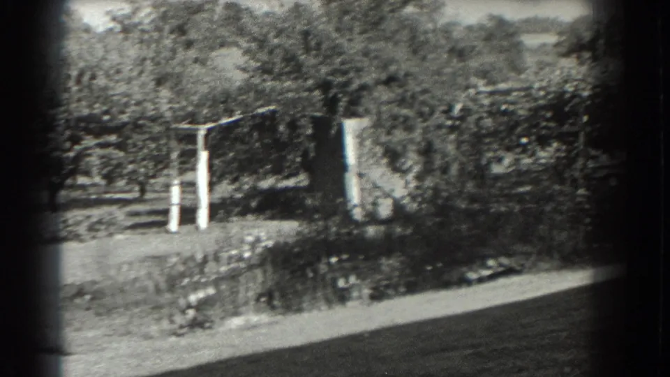 1937 rural Southampton frame with garden beds, a pale wooden gate or support frame, orchard trees, and shrubs beyond the lawn.