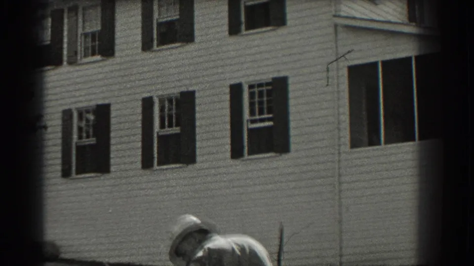 1937 Southampton frame with the house exterior behind a partially seen person in a light hat and clothing near the yard.