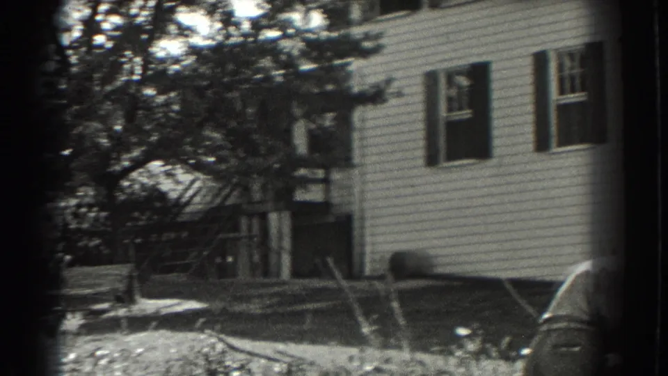 1937 Southampton frame showing the house exterior, trees, simple yard fencing, and the edge of a parked car in the foreground.