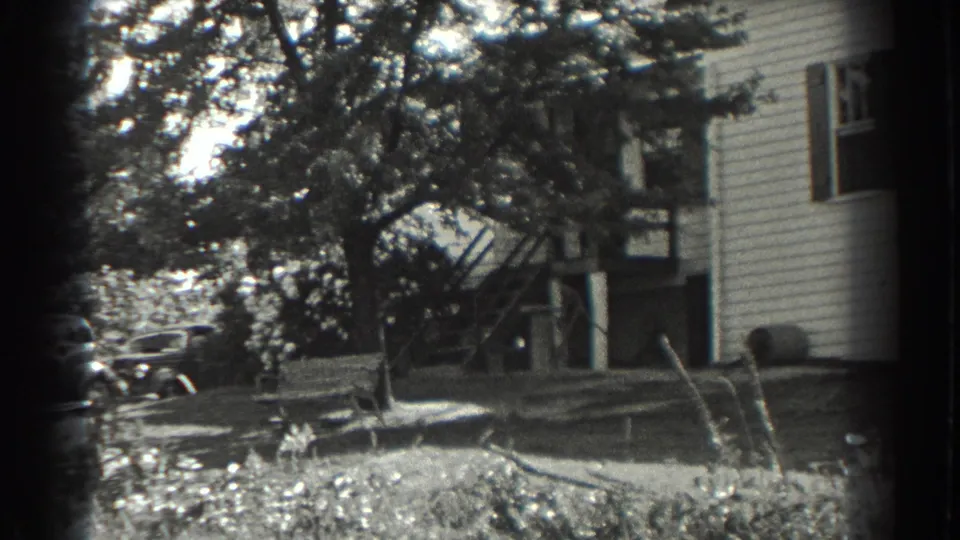 1937 Southampton home-movie frame showing a clapboard house, leafy trees, yard growth, and a partly hidden car near the property.