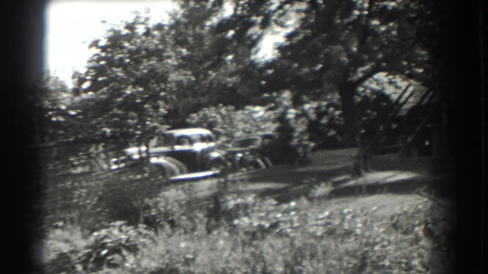 1937 Southampton yard view with a rounded automobile partly obscured by trees, shrubs, and low garden fencing.