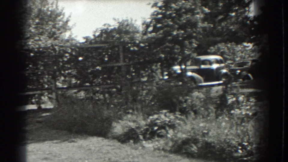 1937 Southampton home-movie frame of a leafy garden area with shrubs, trees, fencing, and a parked car beyond the plants.