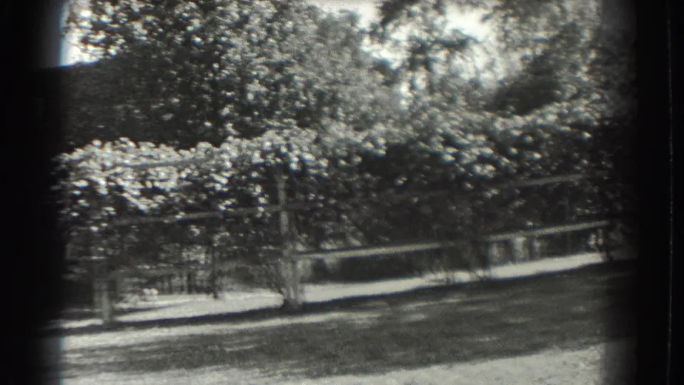 Black-and-white 8mm frame of a Southampton garden with trees, white fencing, lawn, and a trellis-like structure.