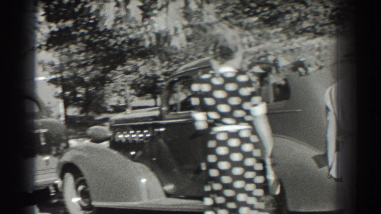 Woman in a polka-dot dress walks beside a parked car under trees.