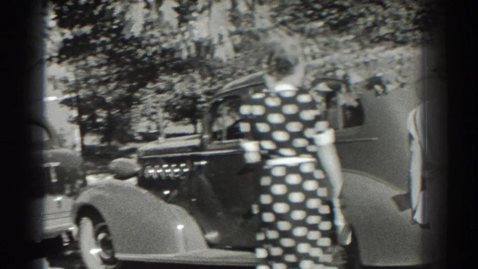 A woman in a polka-dot dress walks beside a classic car in 1937 Southampton, with trees and other vehicles behind her.