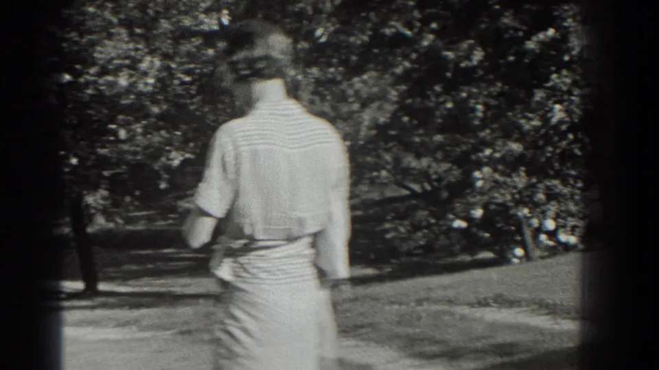 A woman in a light dress and hat walks away from the camera in a leafy Southampton park setting.
