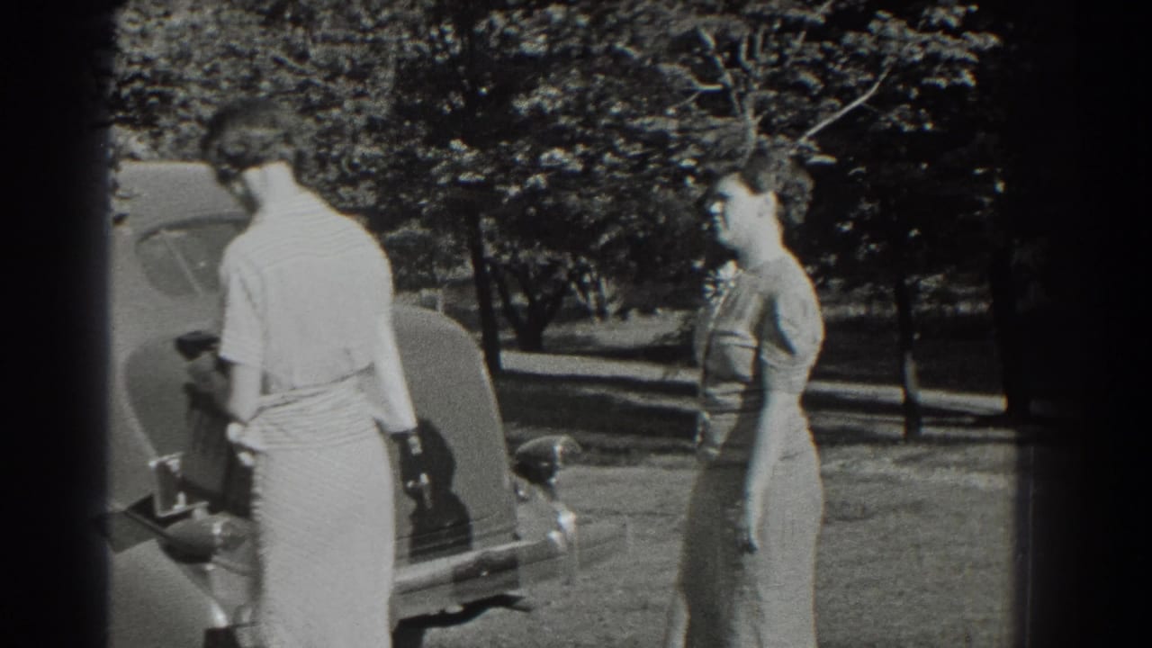 Two women stand near the rear of a classic car in a sunny 1937 Southampton yard.