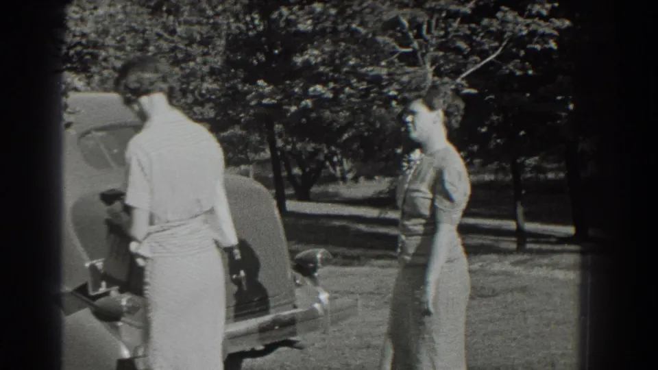 Two women stand near the rear of a classic car in a sunny 1937 Southampton yard.