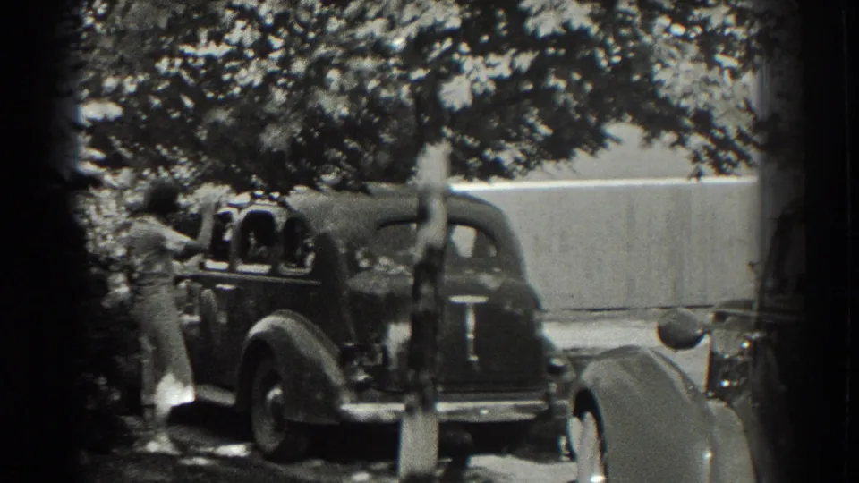 A woman stands beside a parked rounded automobile, framed by trees and a light wall or fence.