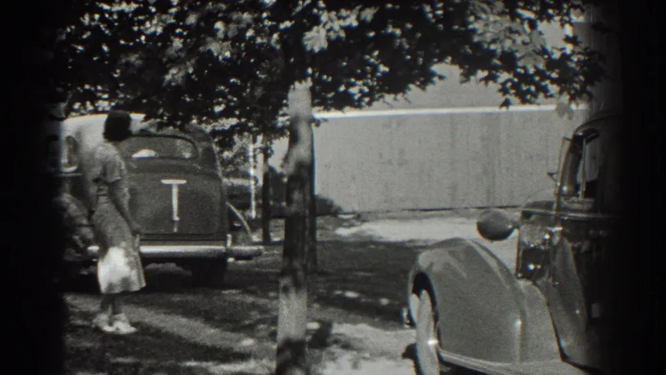 In Southampton, New York, a woman in a light dress walks near parked period cars on a tree-shaded driveway in 1937 home-movie footage.