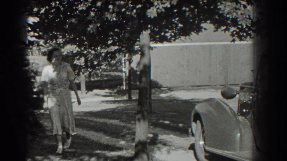 The woman continues across the sunlit and shaded driveway, framed by trees and period automobiles in the 1937 home-movie scene.
