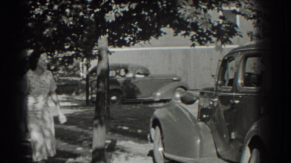 In Southampton, New York, a woman waits beside a tree-lined street while rounded 1930s automobiles sit or pass nearby.