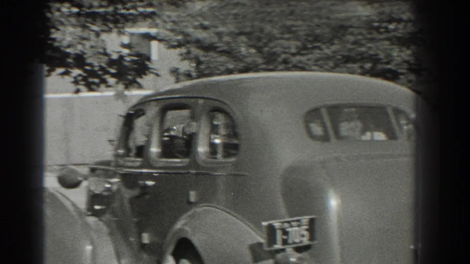 A close view of 1930s sedans on a tree-lined Southampton street shows a rear license plate and soft home-movie focus.