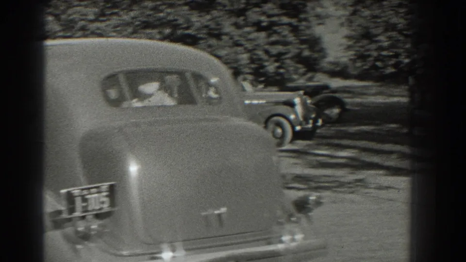 The rear of a 1930s sedan dominates the Southampton street frame, with another automobile crossing the shaded background.
