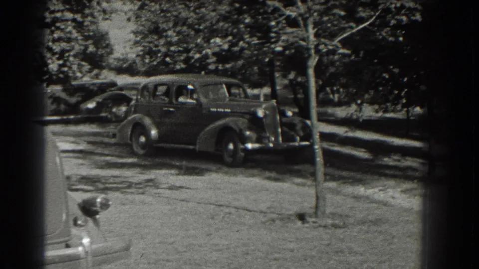 A dark 1930s sedan is seen from the side on a Southampton residential street with trees, shade, and another car in the background.