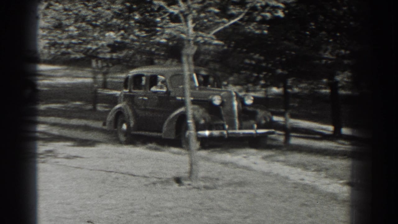 A dark 1930s sedan approaches along a tree-lined driveway.
