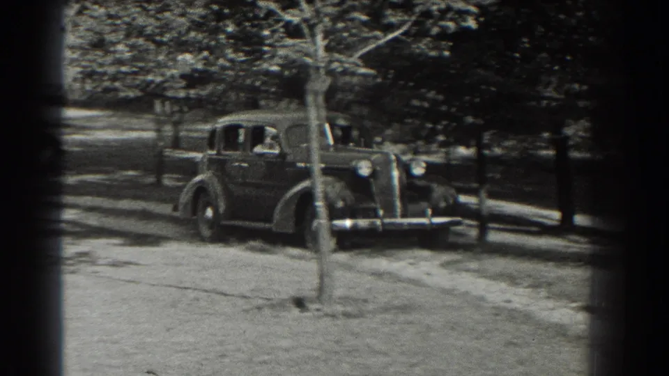 A 1930s sedan approaches along a tree-lined drive in Southhampton, New York, in black-and-white 8mm footage.
