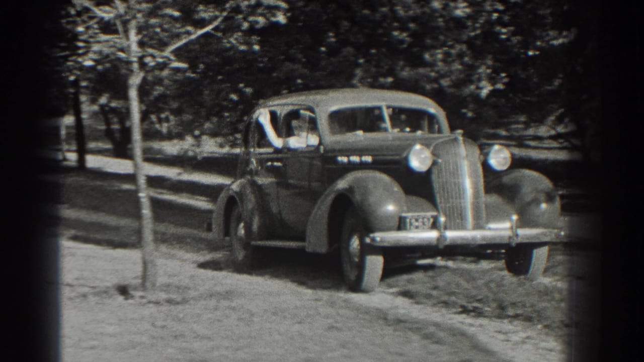The sedan draws closer on the driveway with passengers visible through the windows.