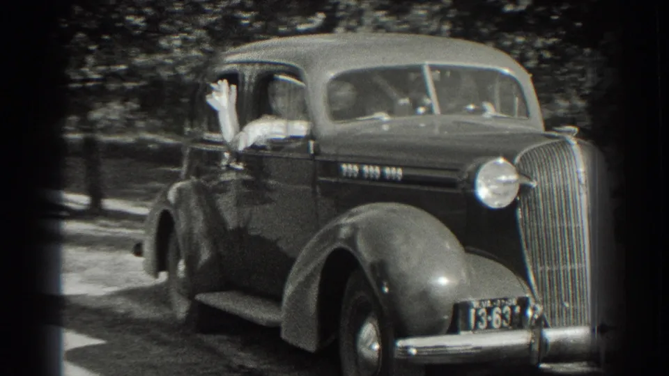 A passenger waves from the side window of a 1930s sedan as it passes close to the camera in Southhampton, New York.