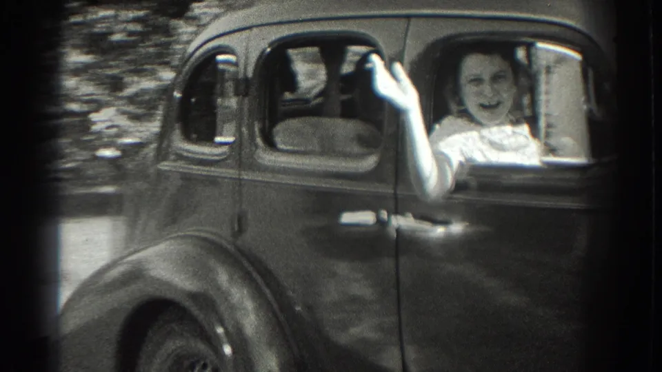 A smiling woman waves from the passenger window of the 1930s sedan, with another occupant visible inside the car.