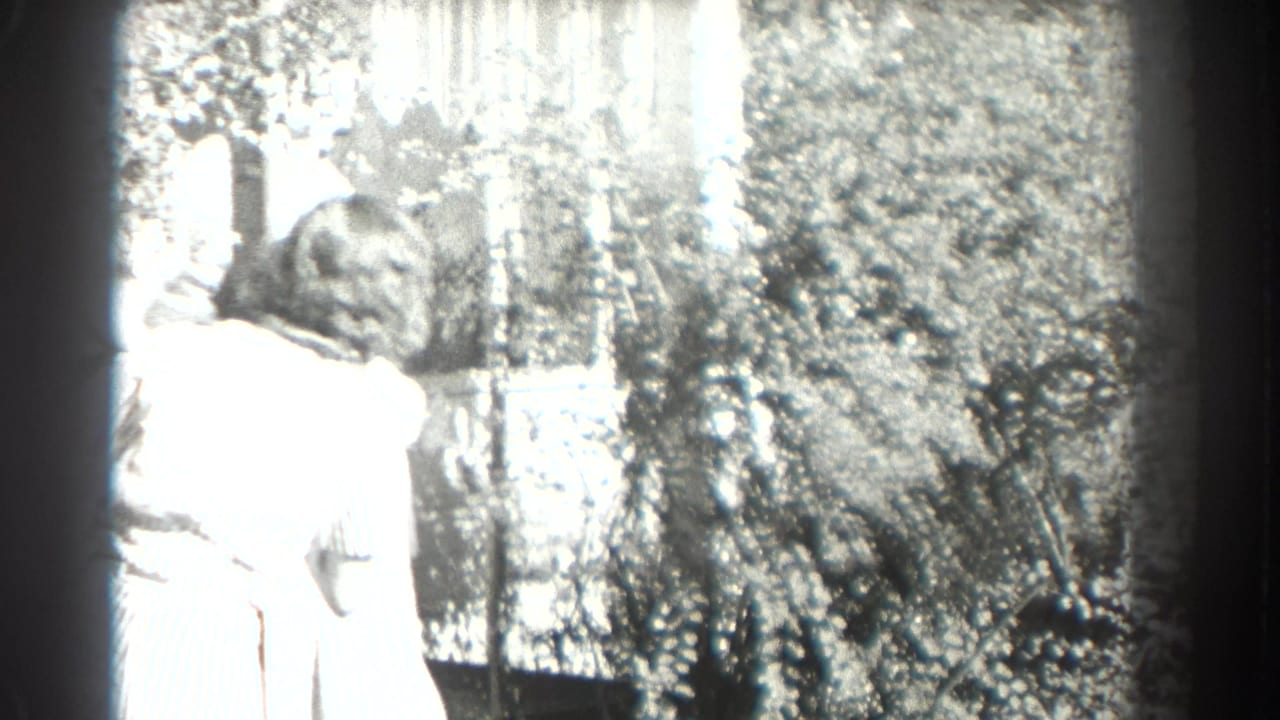 Adult woman in a light dress stands beside a car in a sunny garden.