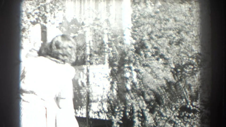 In supplied 1937 Southampton home-movie footage, an adult woman in a light dress stands near a car amid bright garden foliage.