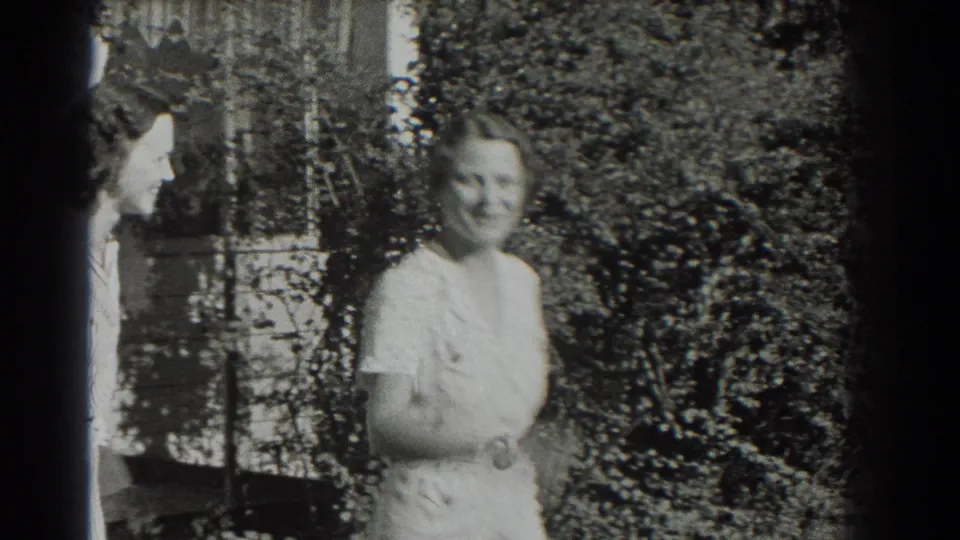 Two adult women in light clothing appear to converse or pose near shrubs and a house exterior in this 1937 Southampton home-movie frame.