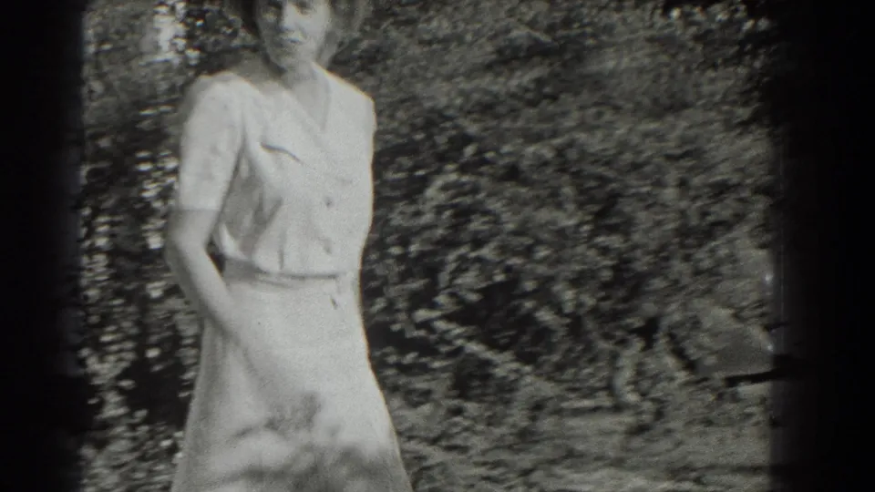 An adult woman in a light dress walks close to the handheld camera in the supplied 1937 Southampton garden footage.