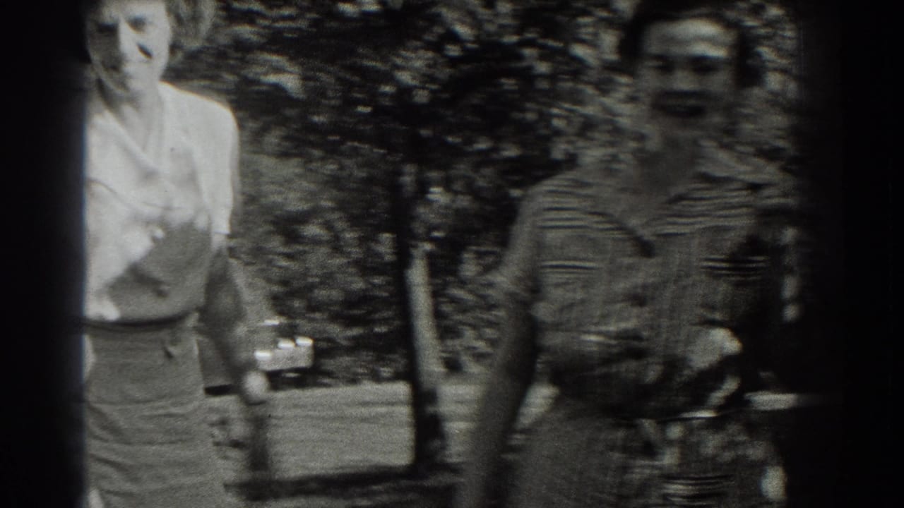 Two women walk toward the camera under trees in black-and-white home-movie footage.