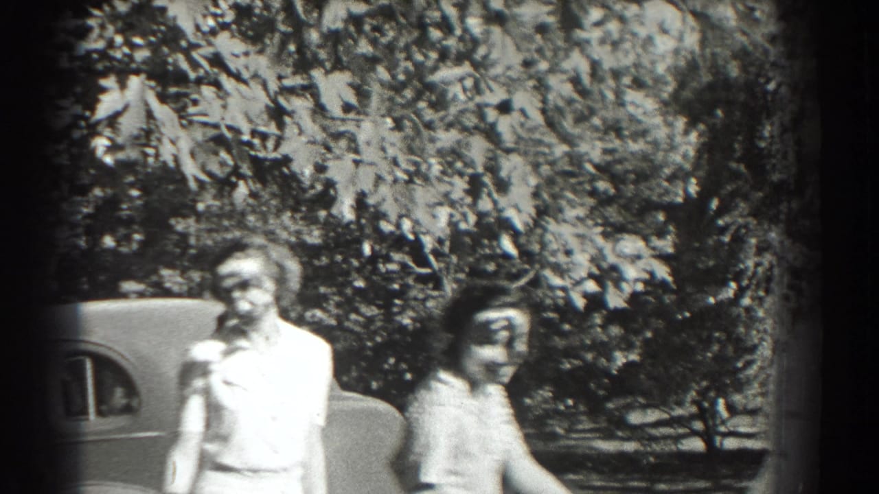 Two women walk past a parked vintage car with trees behind them.