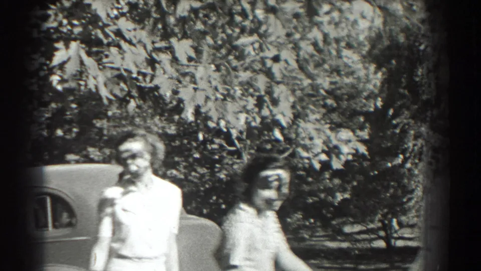 Two women walk through a sunny tree-lined area near a rounded vintage car in 1937 Southampton home-movie footage.