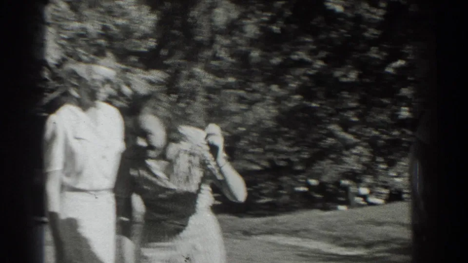 Two women in light and dark clothing appear near dense foliage, captured with soft focus and heavy grain in the 1937 clip.