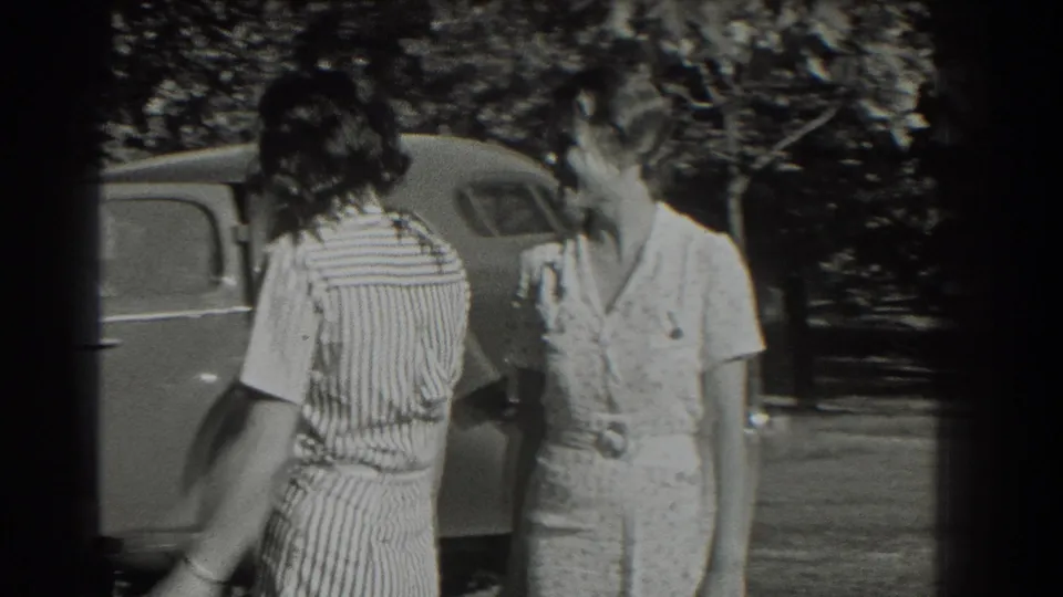 Two young women in summer dresses stand near a parked 1930s-style car in Southhampton, New York home-movie footage.