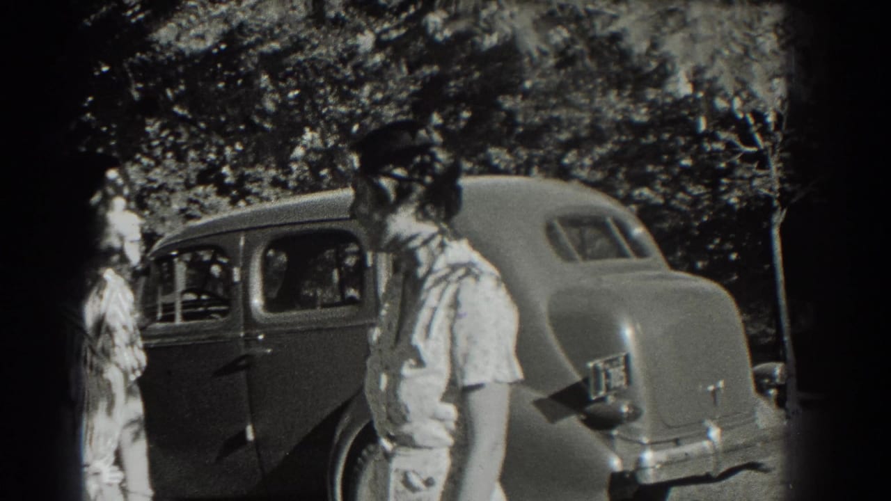 A young woman in a light dress stands near the rear of a 1930s-style sedan, with trees and another person in the background.