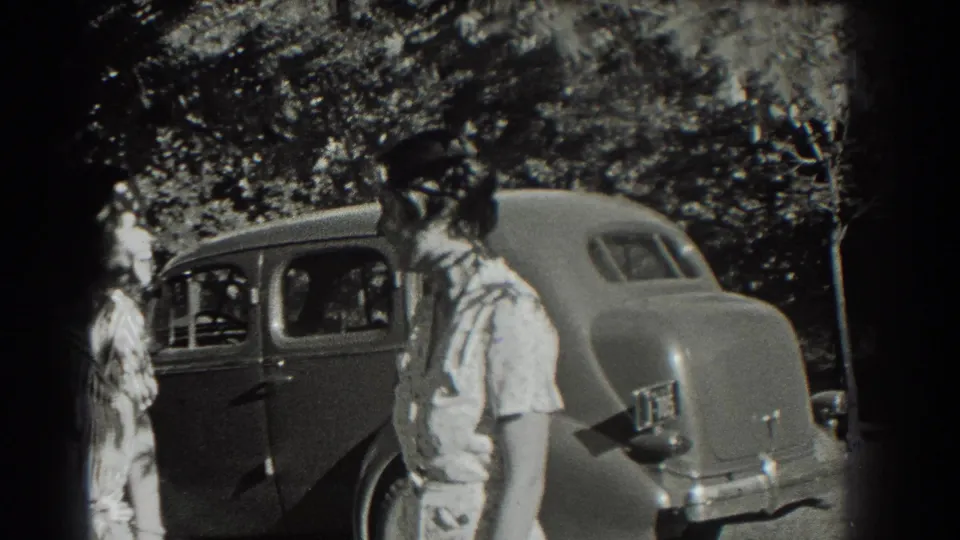 A young woman in a light dress stands near the rear of a 1930s-style sedan, with trees and another person in the background.
