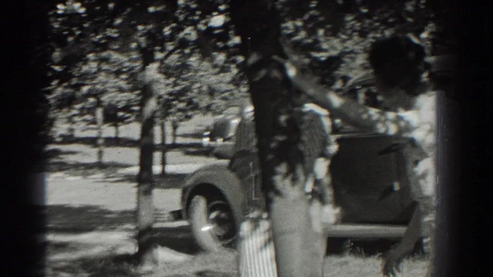 Two young women pass near a tree trunk as the home-movie camera catches a parked car and shaded lawn behind them.