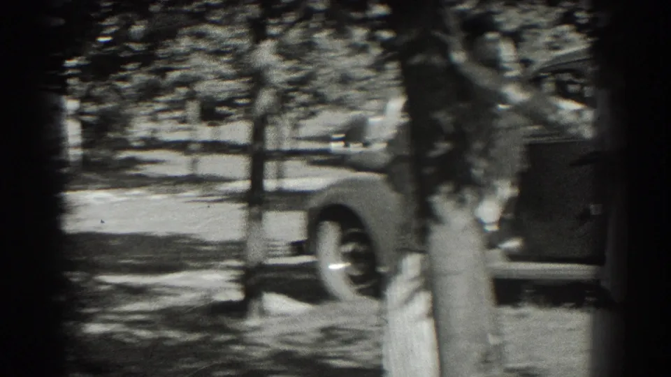Two young women appear near a tree in a shaded outdoor setting, with the parked 1930s-style car partly visible at frame edge.