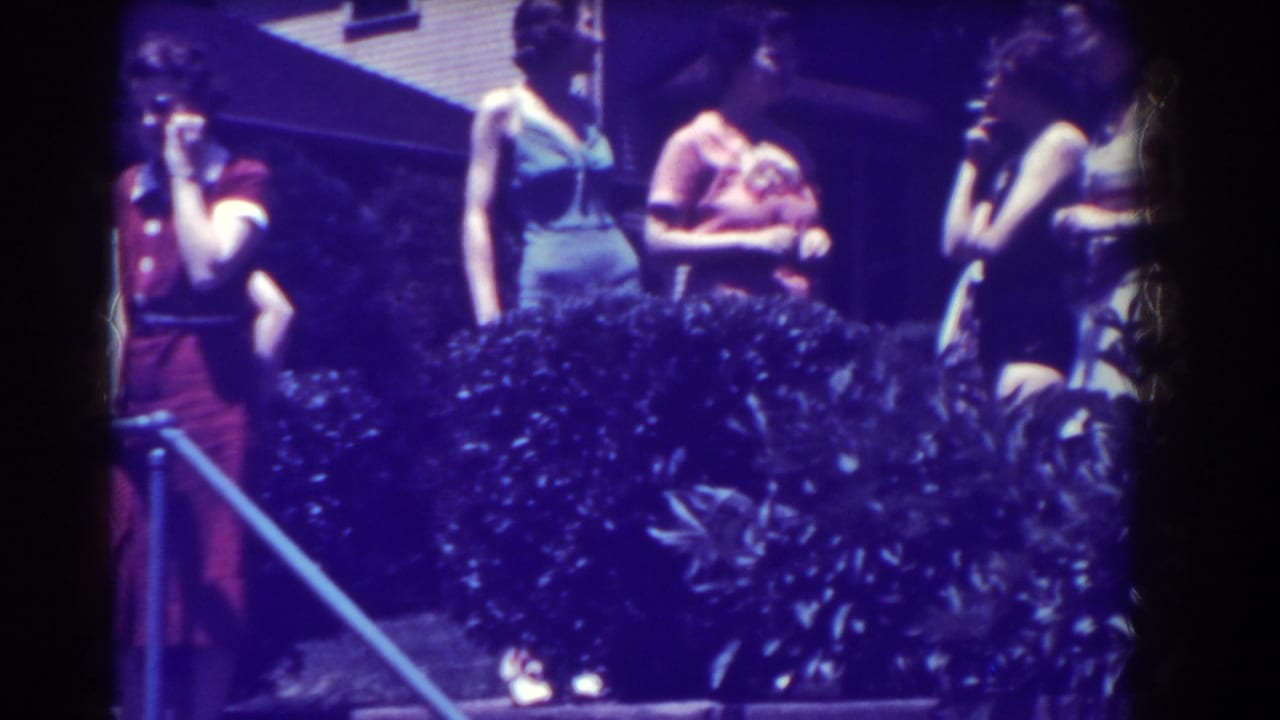 Women in swimsuits stand and talk near bushes beside a pool.