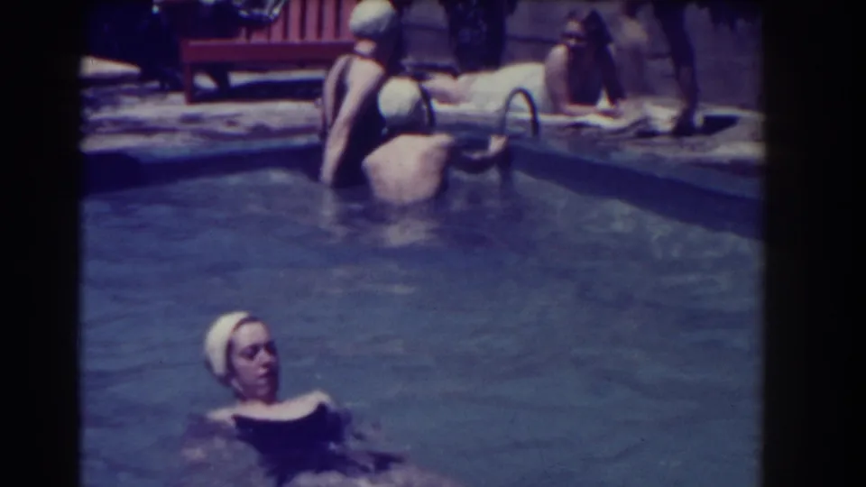 A woman in a white swim cap swims in the pool as other women lounge and stand along the Southampton poolside.