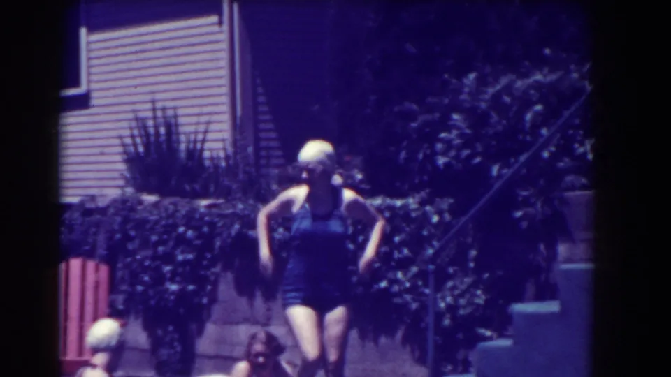 A woman in a 1930s-style one-piece swimsuit walks near steps and plantings by the pool.