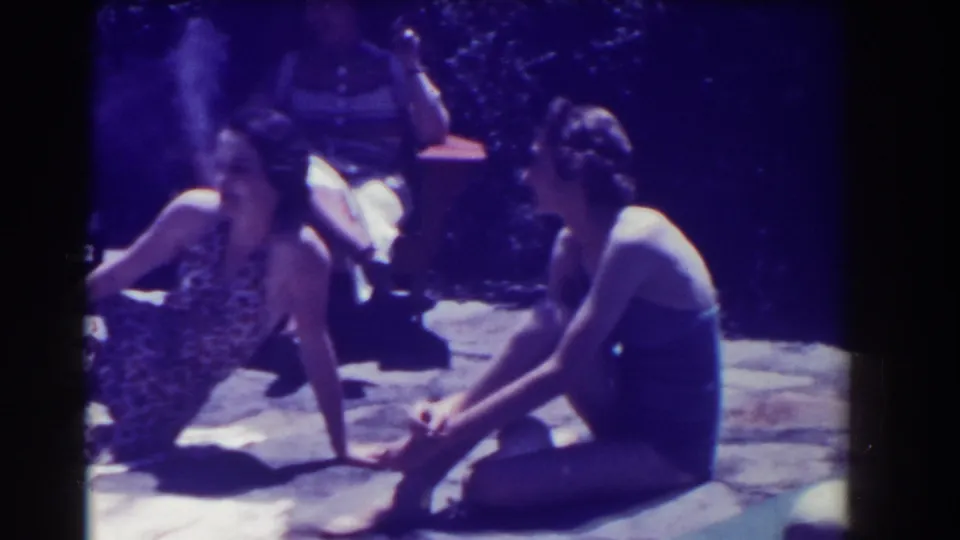 Two women sit and talk outdoors in 1937 Southampton footage, with towels and shaded foliage behind them.