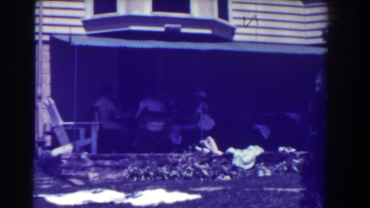 Women seated in shade near a porch and garden outside a house.