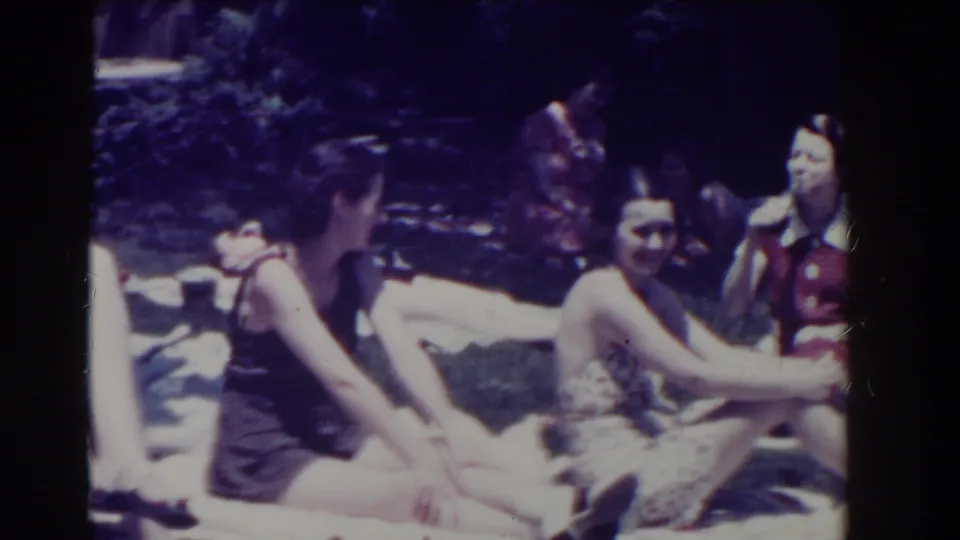 Several women sit together on towels in a sunlit 1937 Southampton garden, with another figure standing behind them.