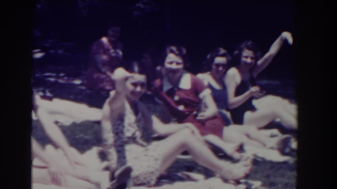 Four women sit on white towels outdoors, with one waving toward the camera.