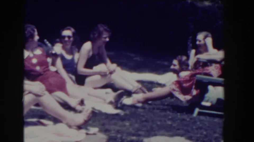 Women relax on towels and appear to talk together outdoors, with a reclining chair visible at the edge of the frame.