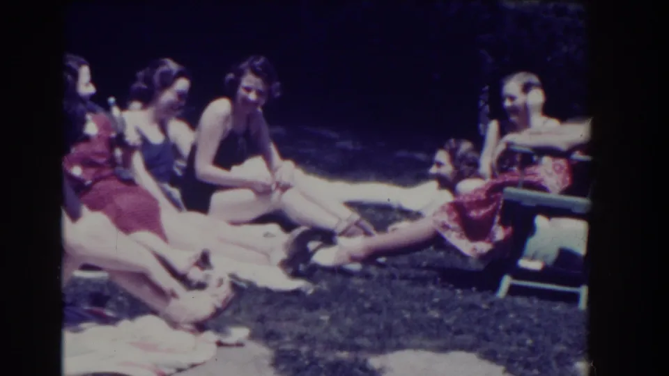 Women in summer clothing sit and recline on towels in a shaded lawn setting, continuing an informal social moment.